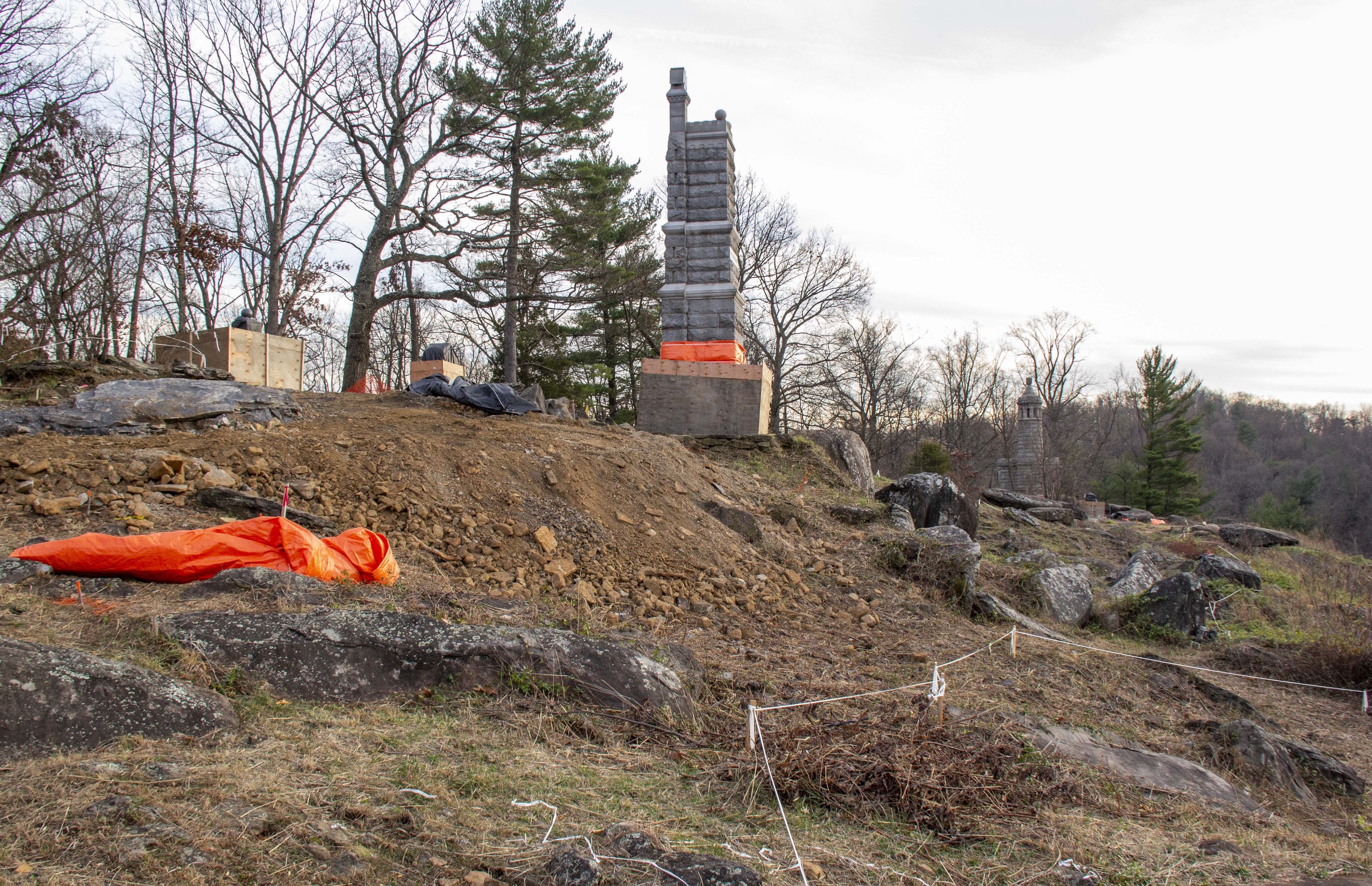 A tall stone monument sits to the right protected by plywood boards at its base and trees and boulders sit to its left. Various construction stakes and white tape mark off areas in front of it.