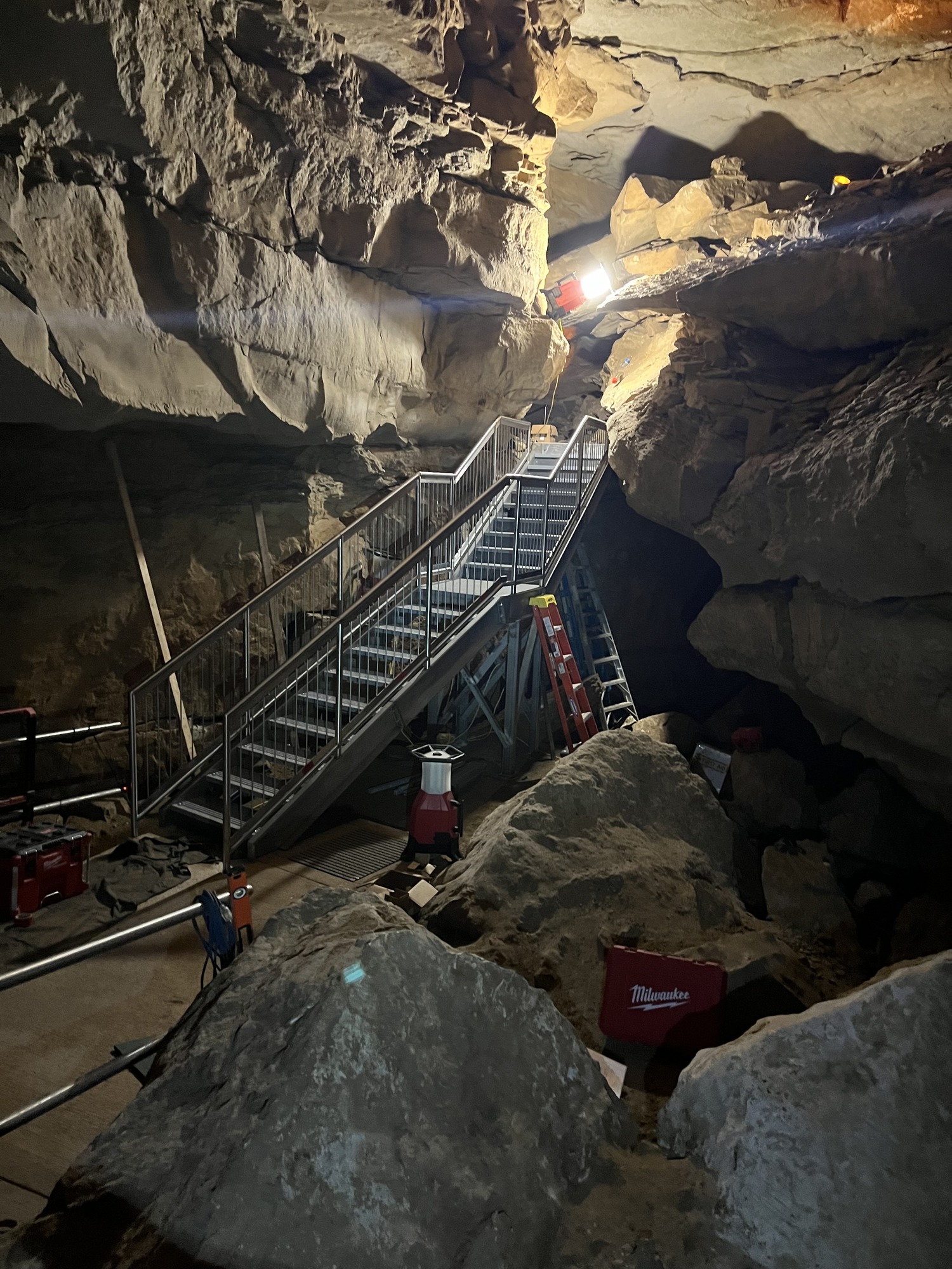 A metal stairway with railings ascends into an upper passage of a rocky cave.
