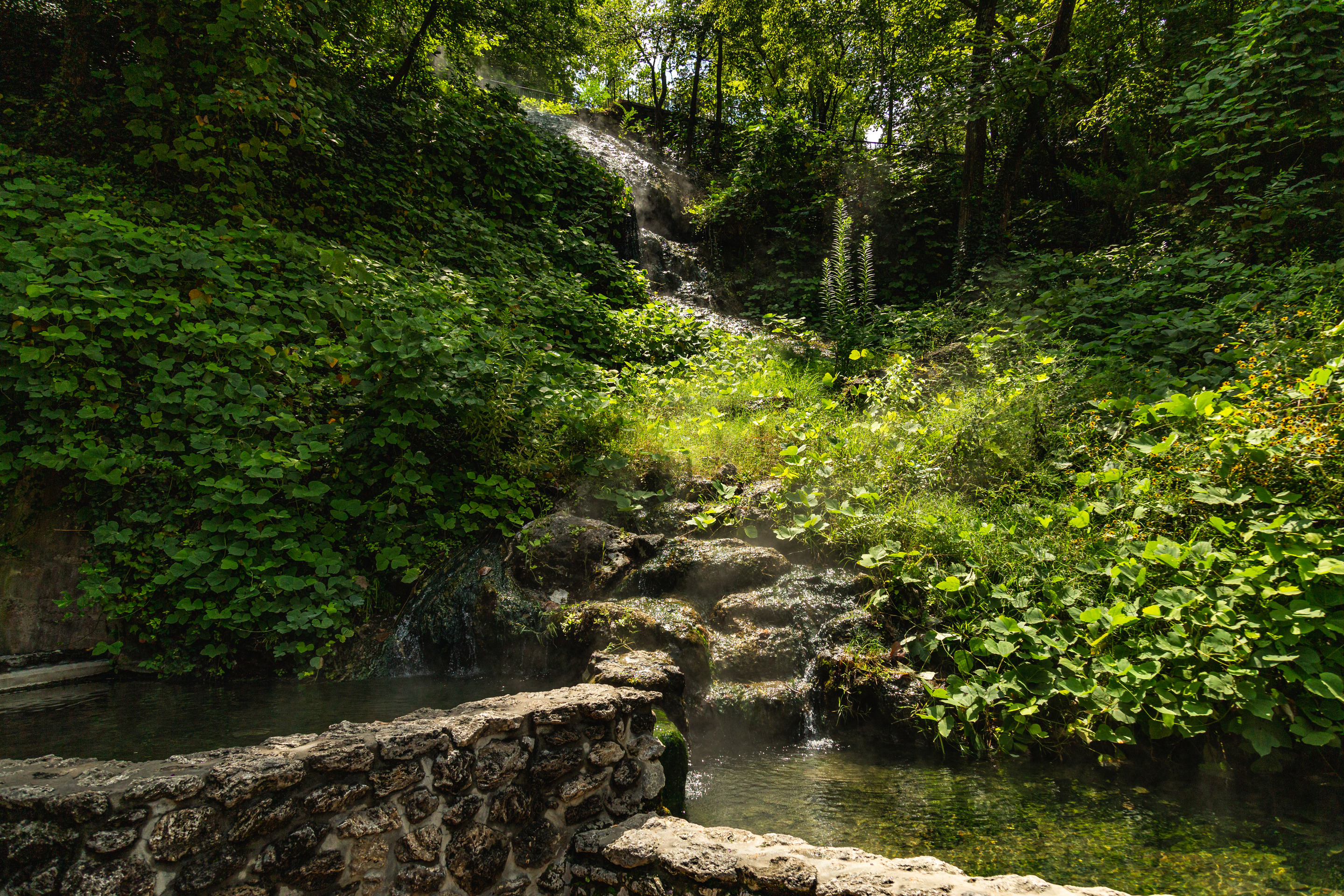 Thermal water spills into two stone-pined pools. The water is traveling downhill through lush vegetation before it reaches the pools.