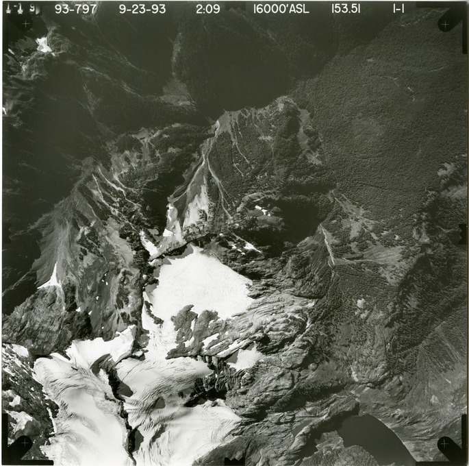 Black and white aerial photograph of glacier on mountain peak, surrounded by forested valleys.