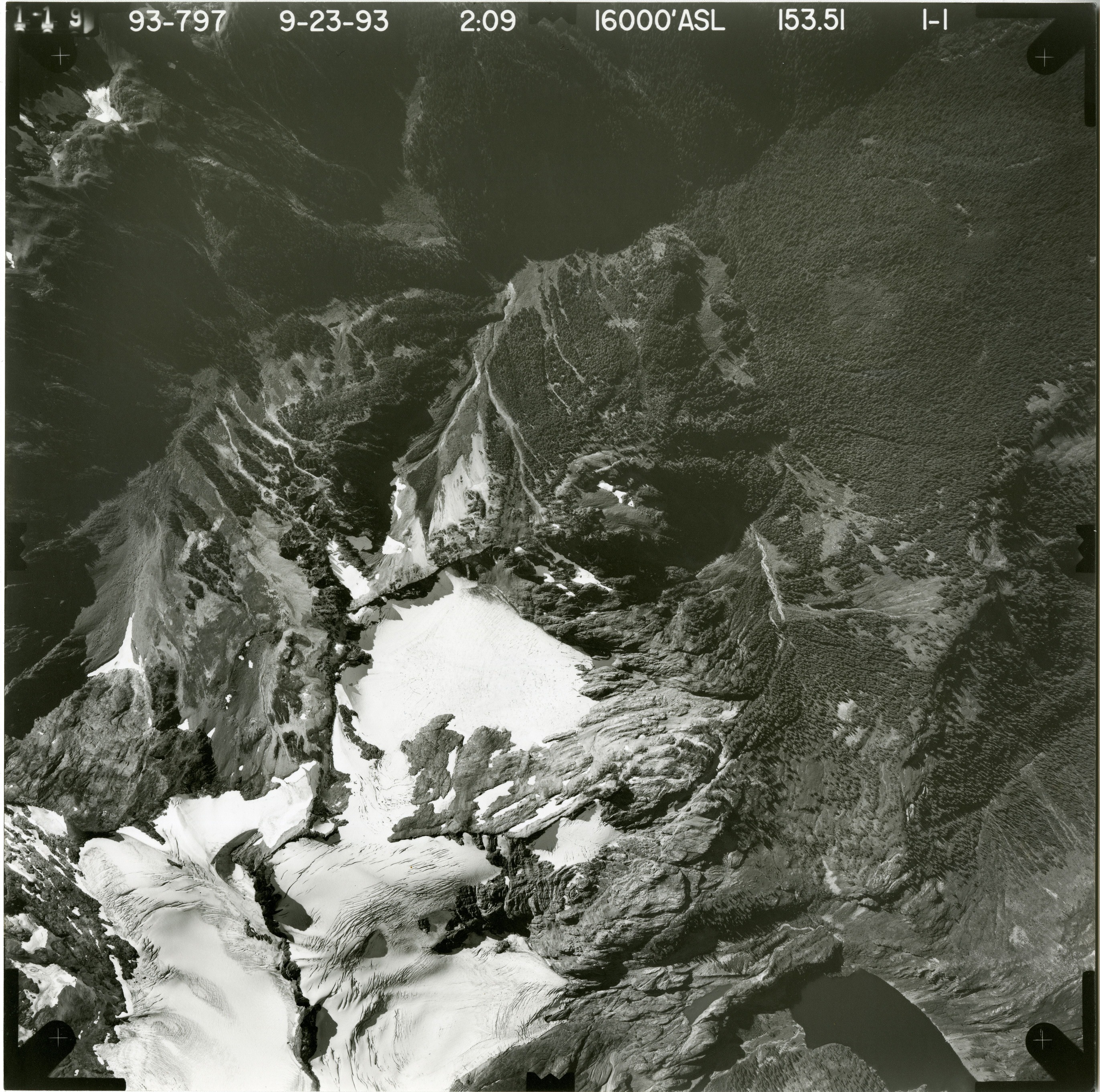 Black and white aerial photograph of glacier on mountain peak, surrounded by forested valleys.