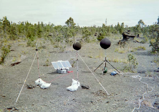 Two spherical microphones sitting on tripods next to a case of equipment, a third tripod with a meteorological sensor, and a solar panel, situated on recent lava rocks with young trees and shrubs.