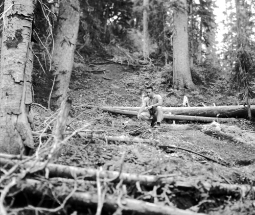 Man with camera kneels next to water supply hose on forest floor. Record of water exploration.