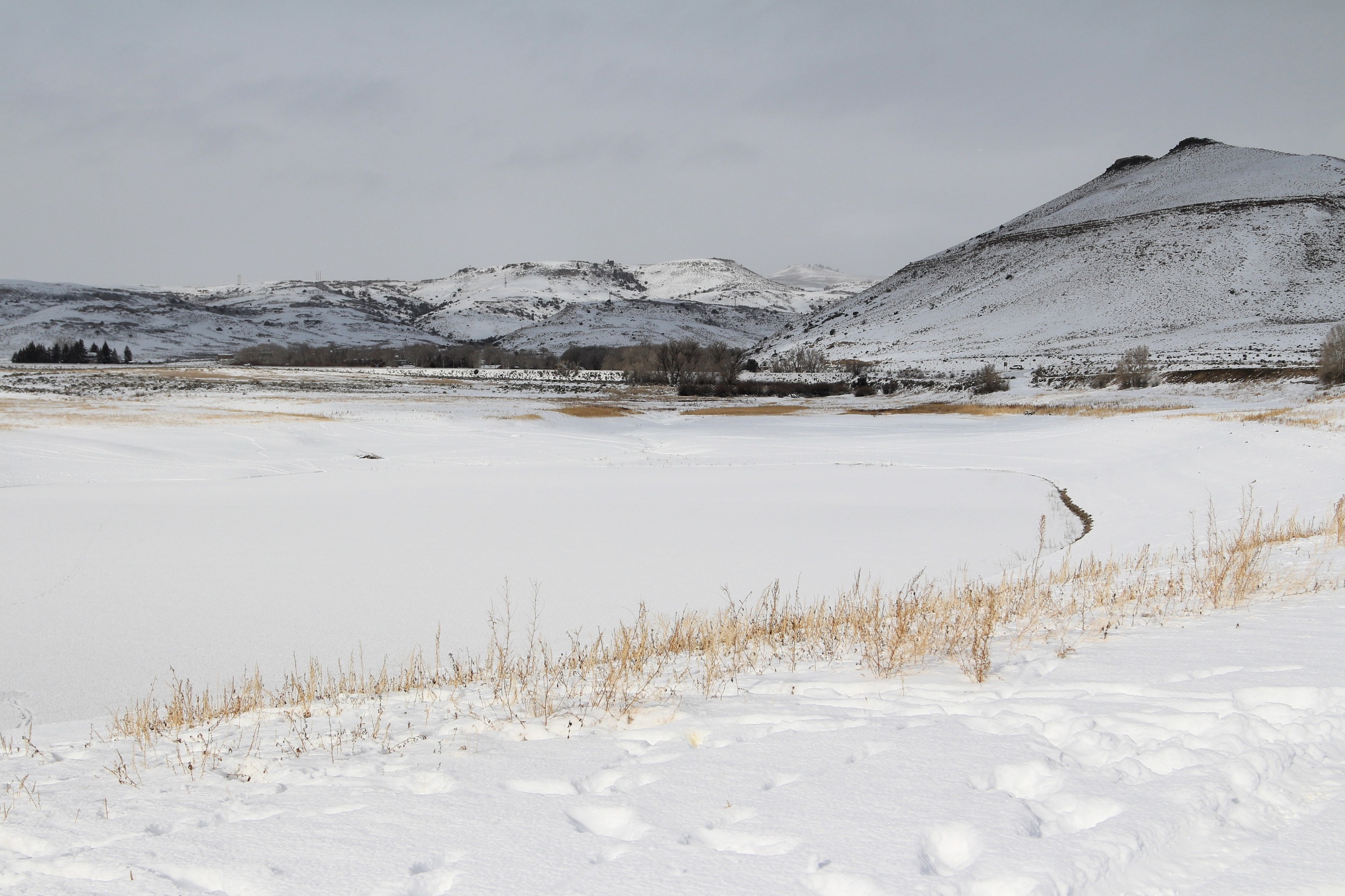 Snow covers a frozen body of water surrounded by mesas. The shoreline has yellow-brown grass and snow with footprints.