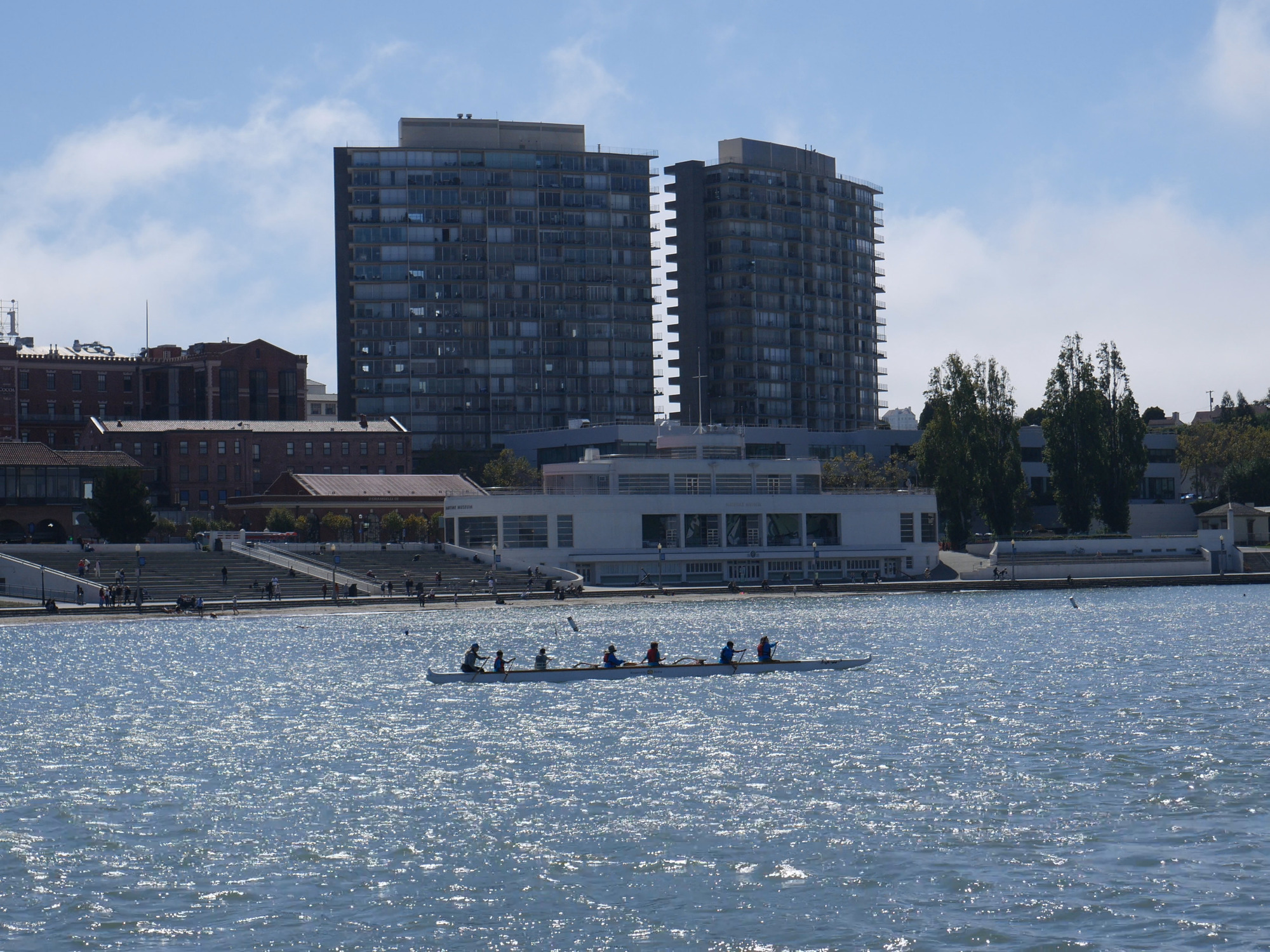 Rowers paddle a long rowing boat in the cove, with the Maritime Museum in the background