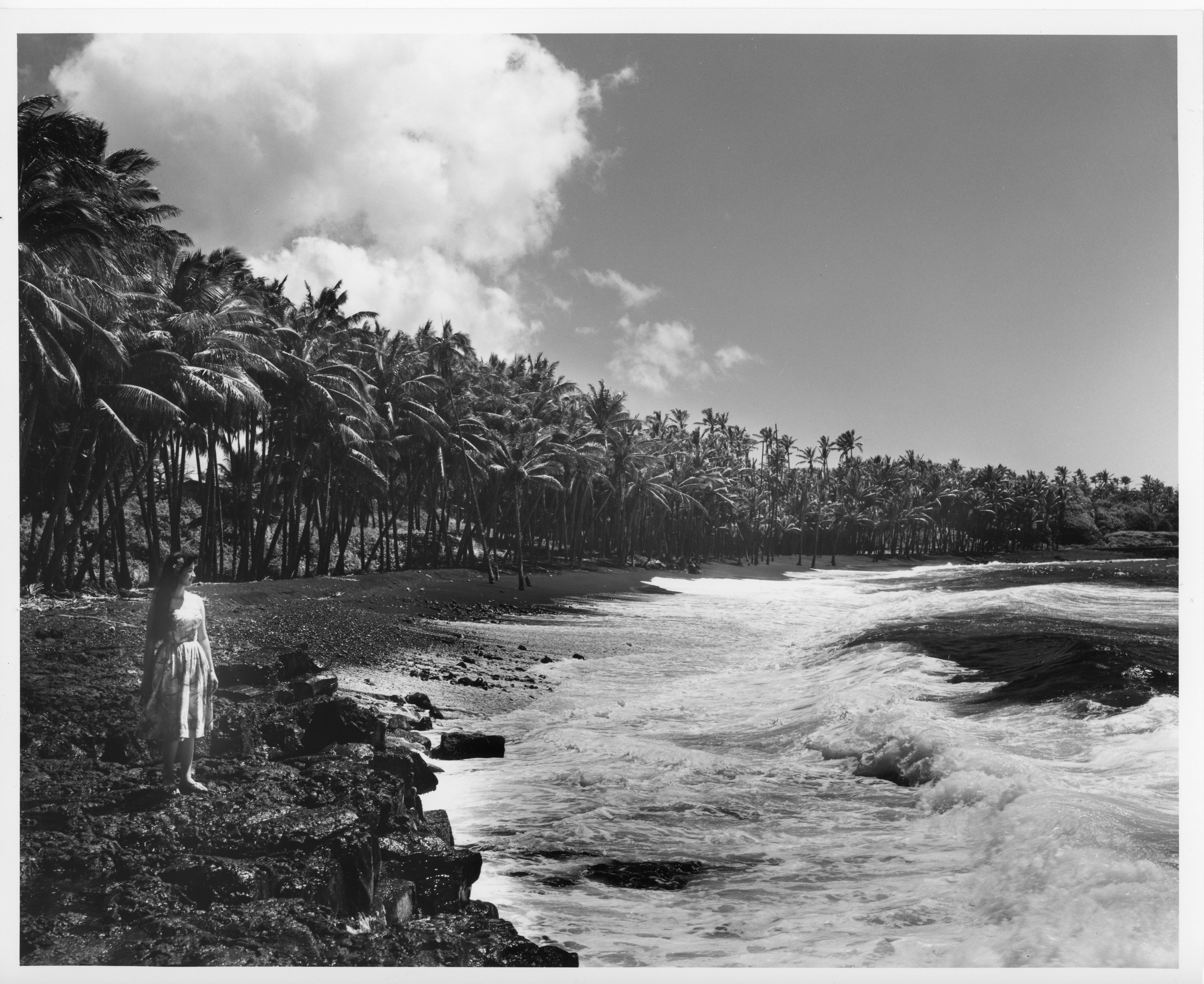 A black and white image of Kalapana black sand beach in Puna, Hawaiʻi.  The image shows the ocean current coming into the shore of the beach which can be seen on the right side of the picture. There is a woman standing on the lava rocks near the water wearing an aloha dress with a flower in her long hair.  She is located on the left side of the image. She is looking out to towards the right side of the image. The black sand and lava rocks are located on the left side of the image. The rocks and black sand are bordered by a line of palm trees and other trees.