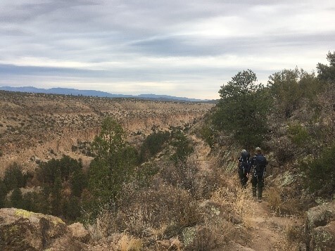 Two hikers head down a rugged trail that leads into a canyon. Blue mountains are far-off in the distance. 