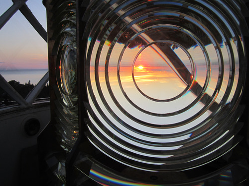 A circular glass lighthouse lens at the top of a lighthouse tower shows the vibrant orange and pink sunset through the center glass. 