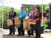 Four colorfully dressed men with drums on shoulder straps underneath a sun canopy, trees in the background.