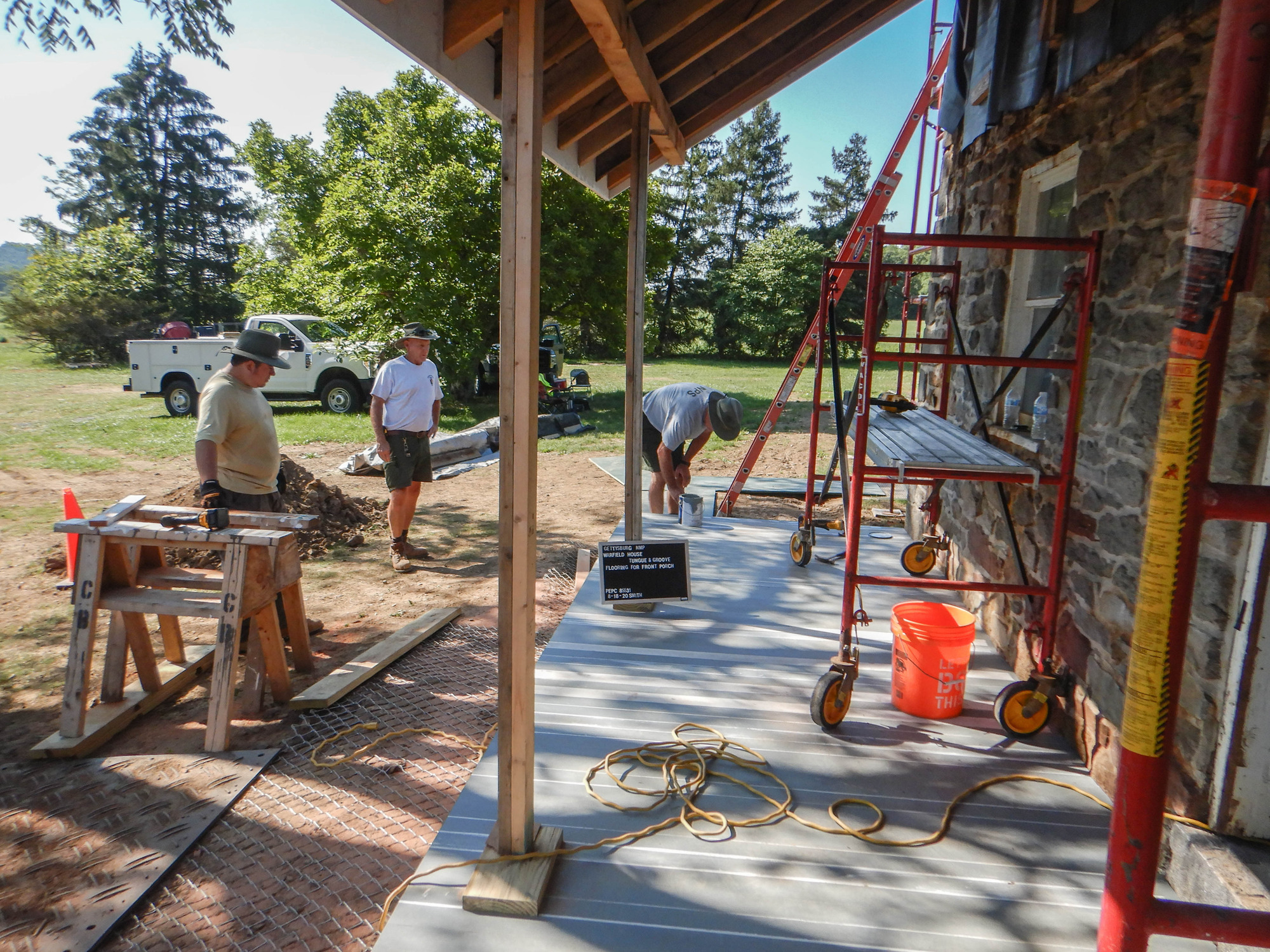 Three members of the preservation crew stand around the porch. There are red scaffolds on the porch and an orange ladder propped against the front of the house. One crew member leans over the porch next to a can of gray paint. There is a white truck in the front yard and various construction equipment and green trees around it. A black and white sign leans against a porch post that reads "Gettysburg NMP Warfield House Flooring For Front Porch PEPC 85531 8-18-20 Smith." 