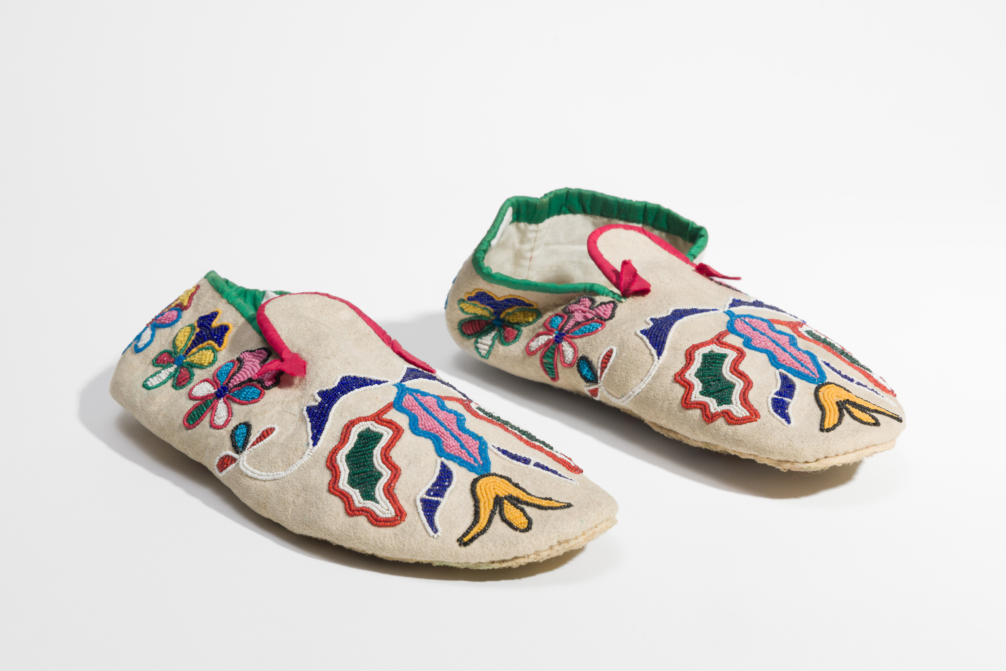 Pair of off-white deerskin moccasins, bound along the top edges with red and green cloth tape and decorated with multi-colored beads in stylized floral design all over the shoe
