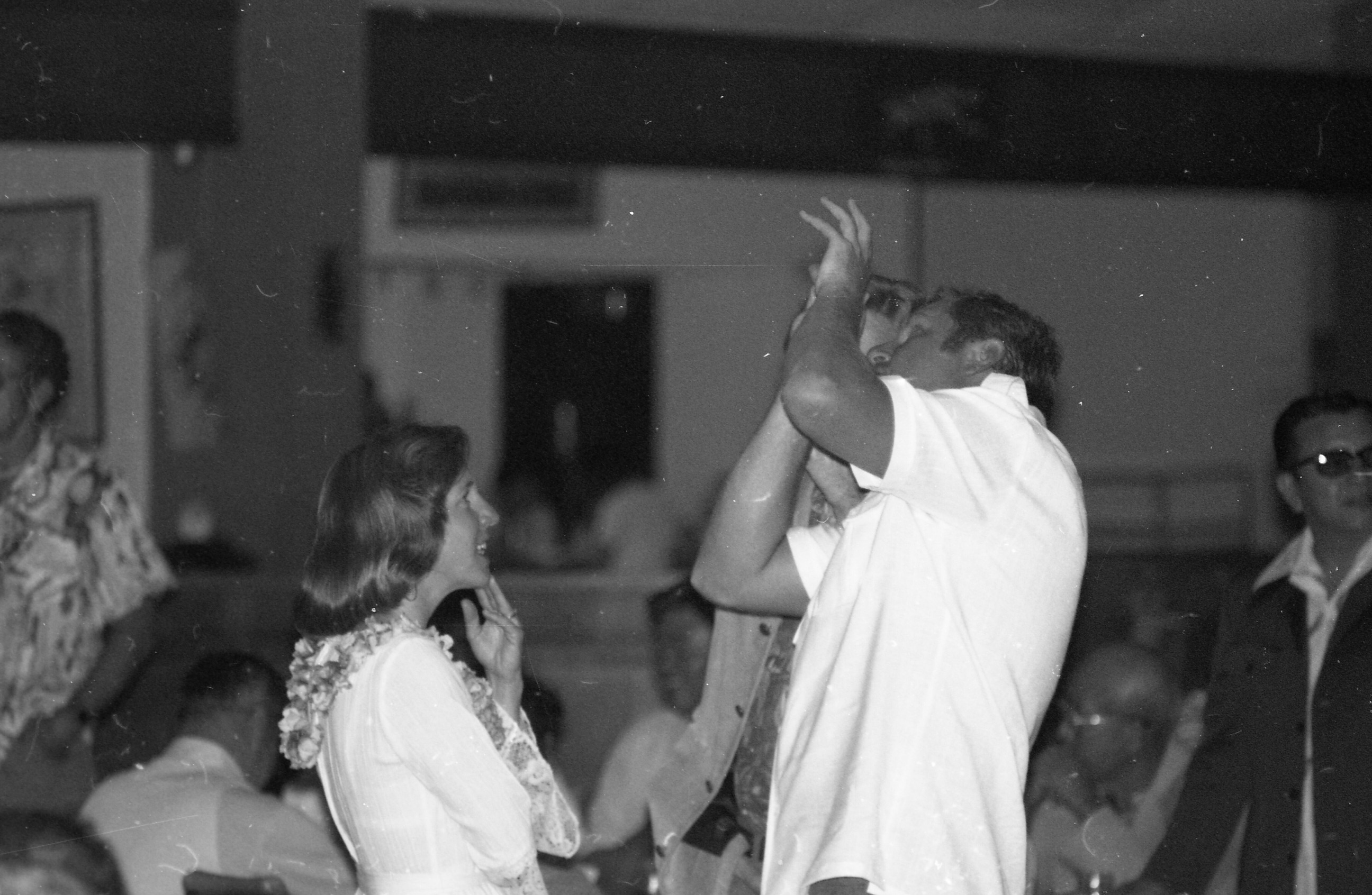 A black and white image of a man and woman conversing during an event. The man and woman are turned facing towards each other unaware a photo is being taken. The woman is located towards the left side of the image. She is wearing a lei and a light-colored long sleeve shirt. Her left hand is touching her chin as she smiles up at the man in front of her. The man is located towards the right side of the image standing in front of the woman. He is very animated using his hands as he talks to the woman. His hands are raised in front of his face leaning slightly backwards. He is wearing a light-colored short sleeve polo shirt. In the background other attendees of the event came be seen siting down at tables and standing around talking to one another.