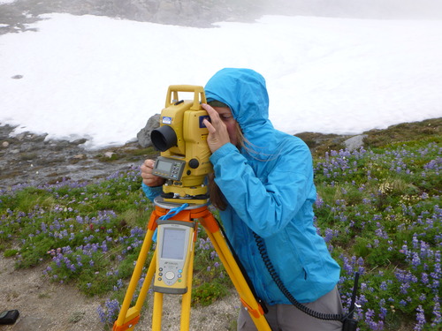 a young woman in a blue rain jacket looks through a survey instrument on a tripod sitting atop a glacier with some purple wildflowers. 