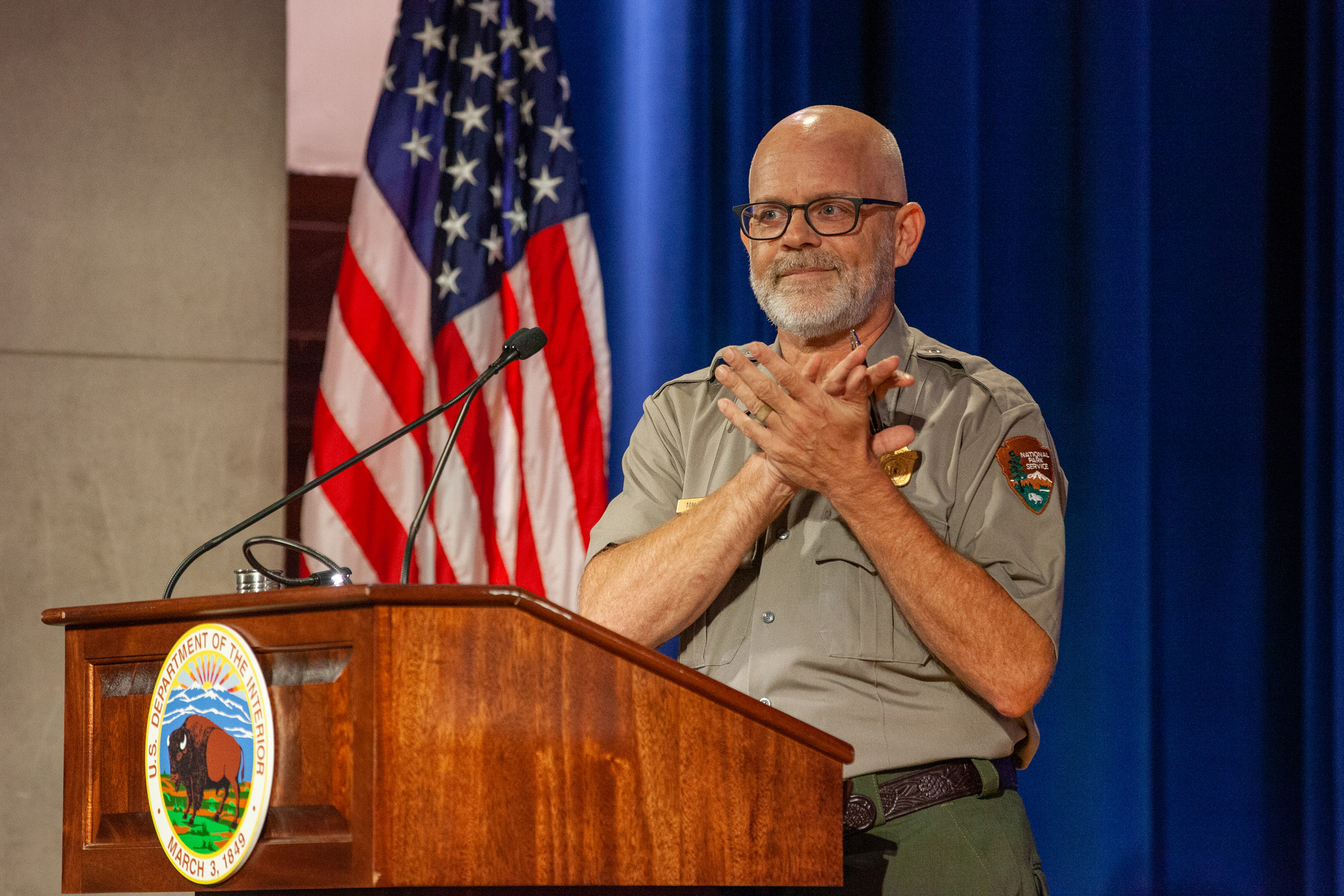 A man in a park ranger uniform and glasses stands clapping at a podium on a stage. 