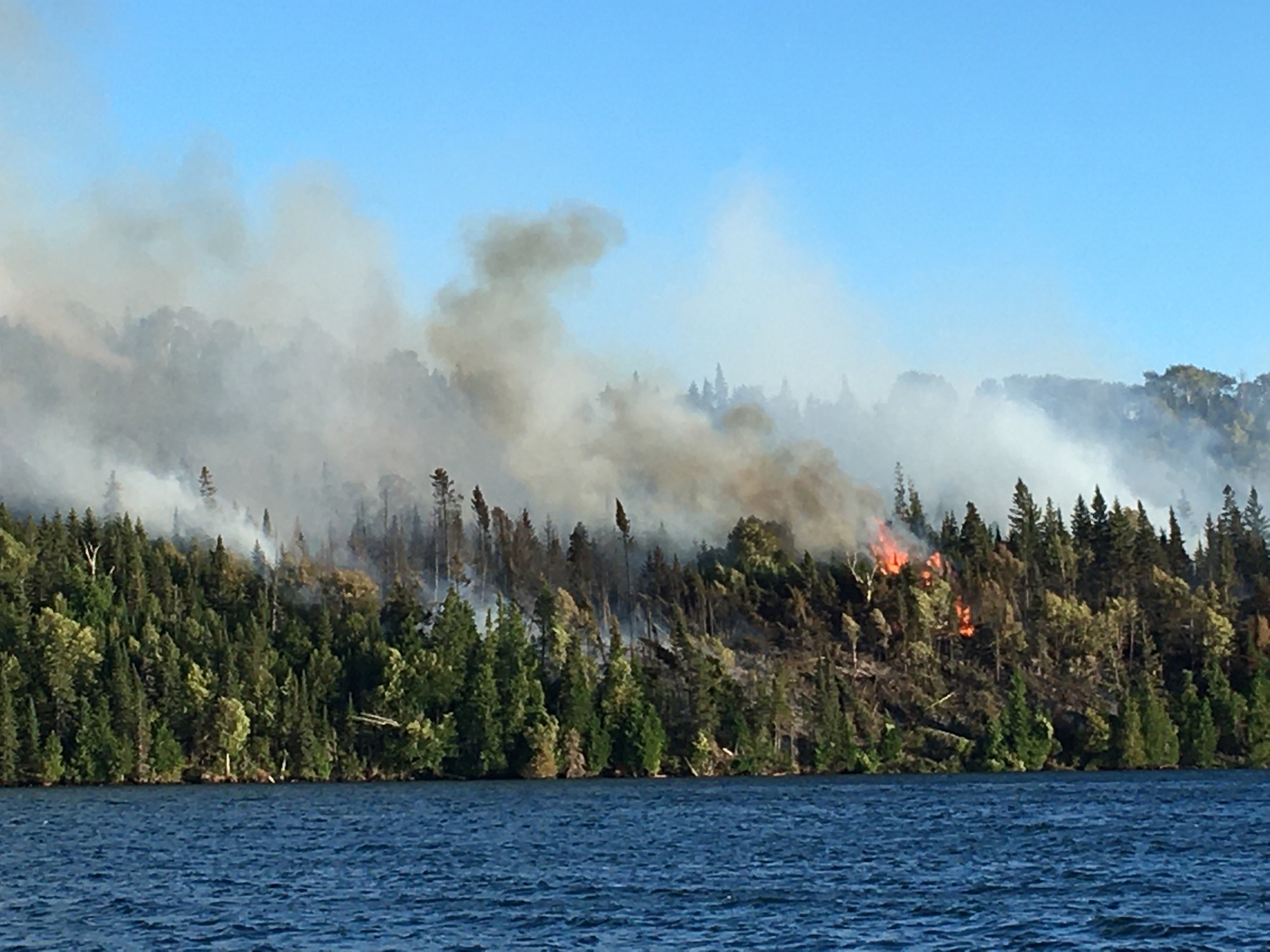 View from the water of a forested shoreline ablaze with bright orange flames and gray smoke billowing into the sky.