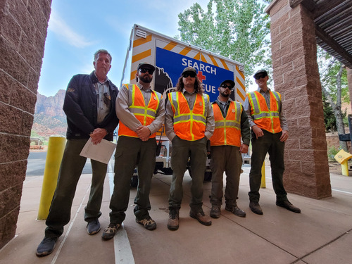 Five rangers pose in front of the search and rescue ambulance. Four of the rangers are wearing orange reflective vests and the last ranger on the left is wearing a jacket over his uniform.