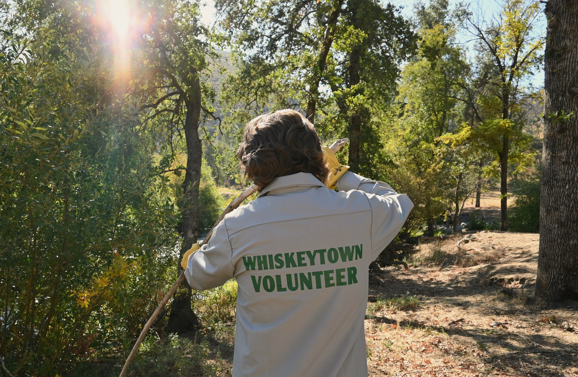A trail crew volunteer picks up debris on a sunny day, surrounded by trees on the trail. 
