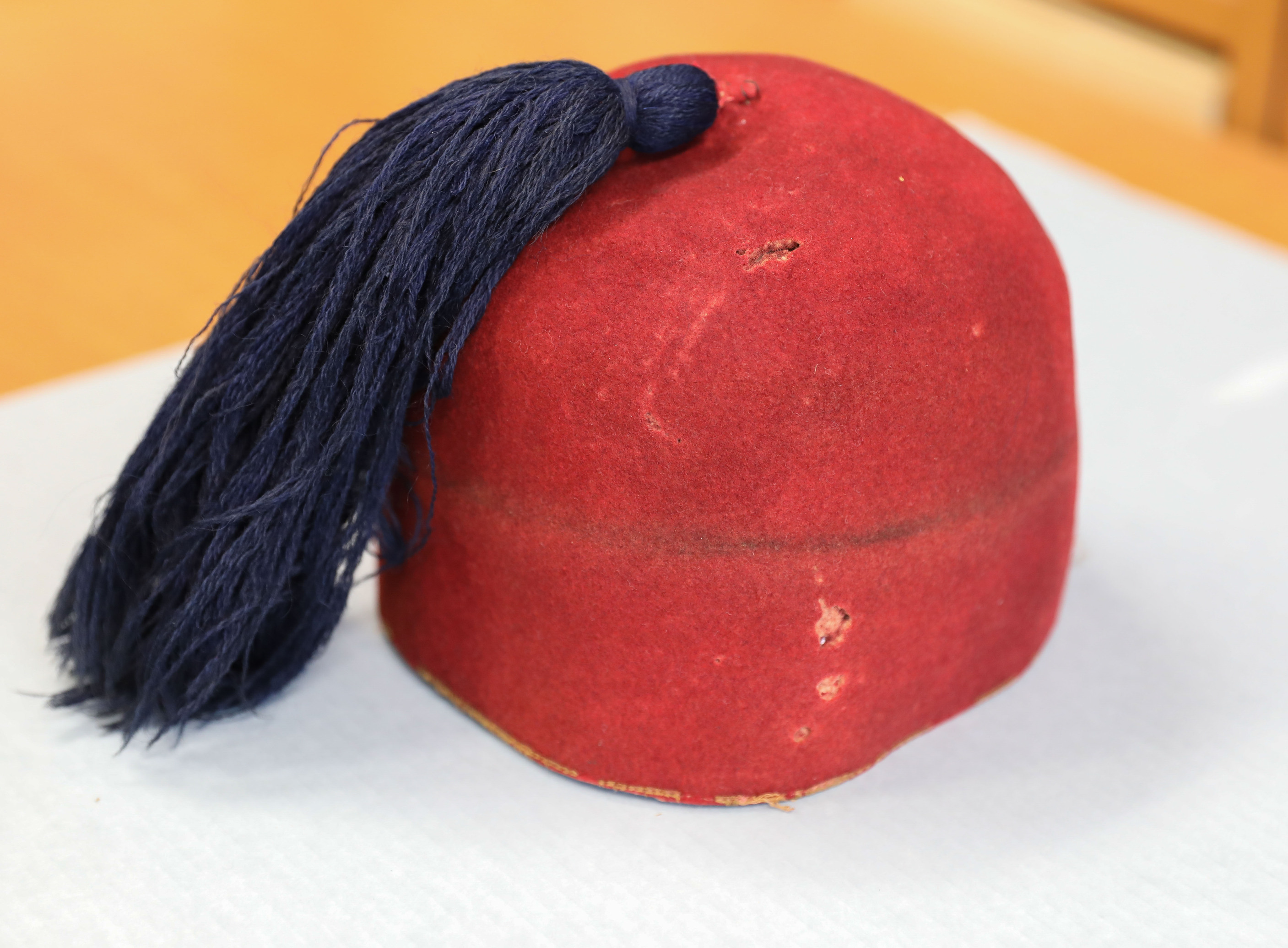 Color photo of the uniform fez of Francis Brownell. The fez, shown from the side, is made from an orange suede-like material and shows some wear and small tears. The tassel that hangs from the middle is made from a navy blue yarn.