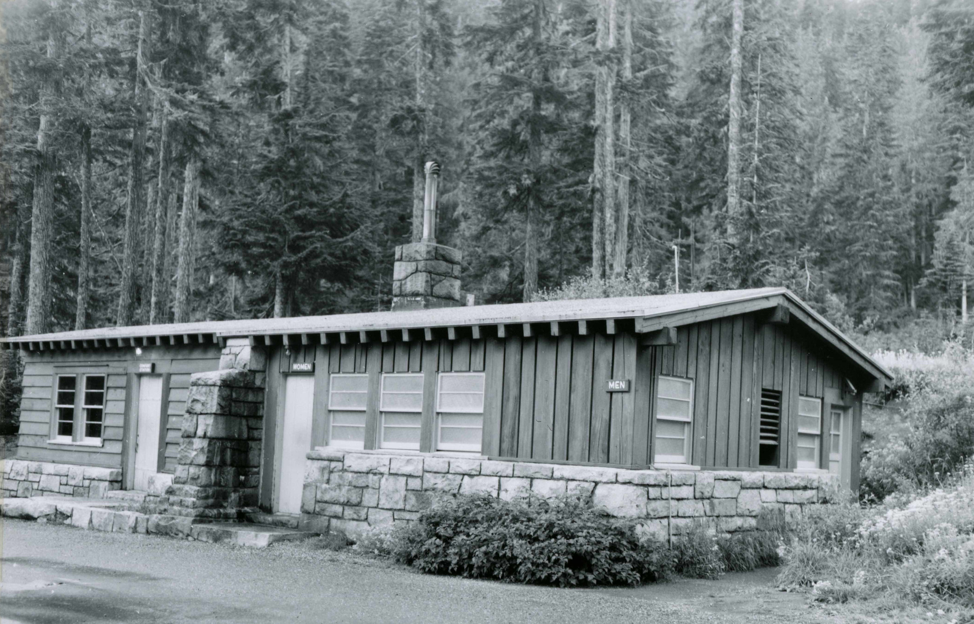 A black and white photo of a small rectangular building with a stone foundation and wood plank sides. The left side has a common room with restrooms on the right, divided by stone wall. 