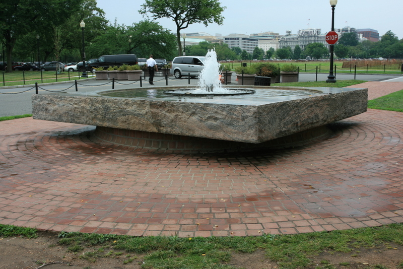 Water sprays up out of the center of fountain that is round pool of water in a stone square base, surrounded by a red brick patio and an urban landscape. 
