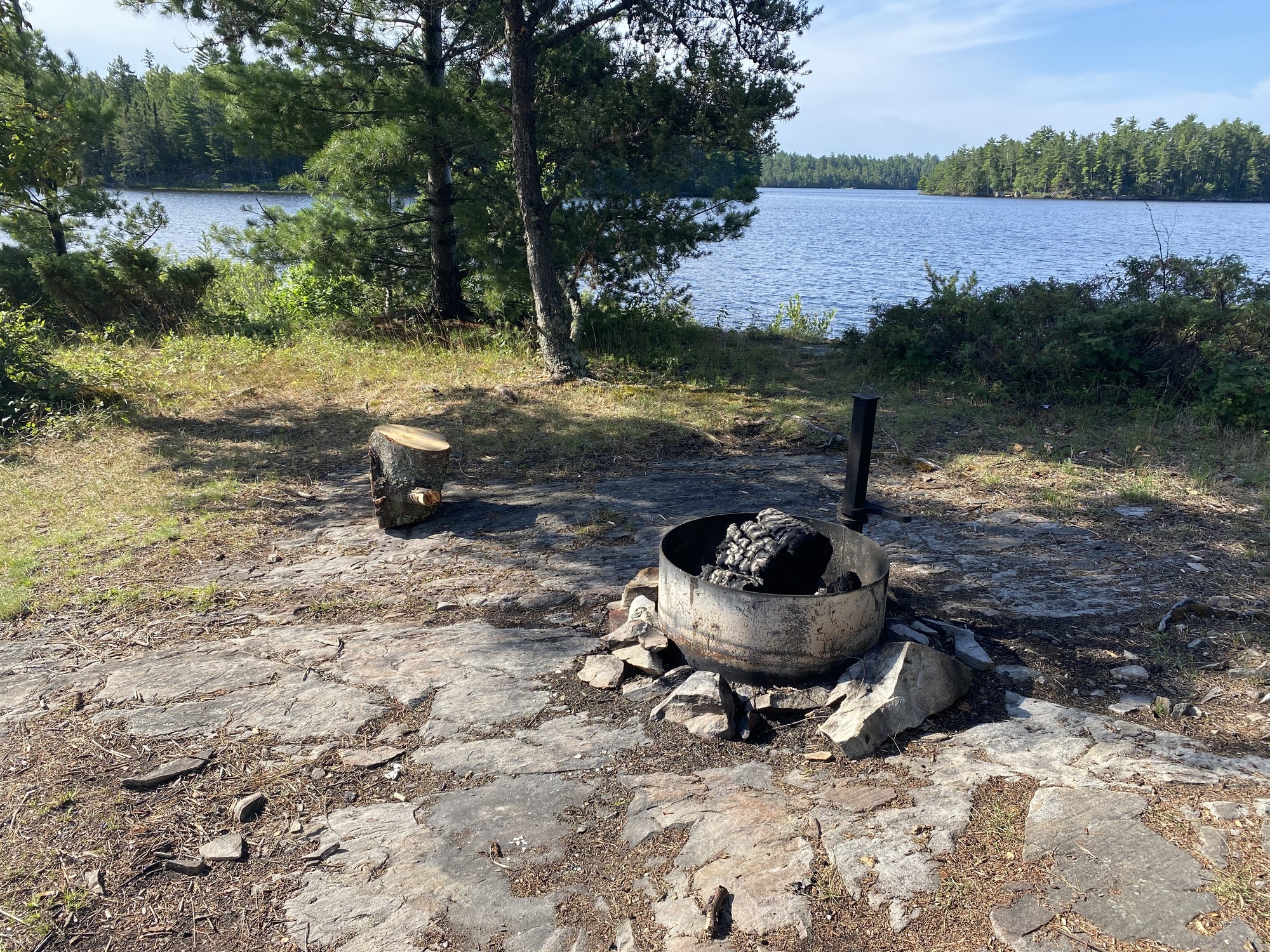 View of houseboat site core and fire ring with a large log next to fire ring. Shrubs and a couple trees border the shoreline with tree covered islands in the background across water.