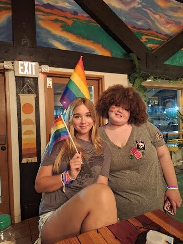 Two young adults in gray shirts pose next to each other at a table holding rainbow pride flags. One person with short curly hair has a rainbow flag on their cheek. Both are wearing colorful bracelets.
