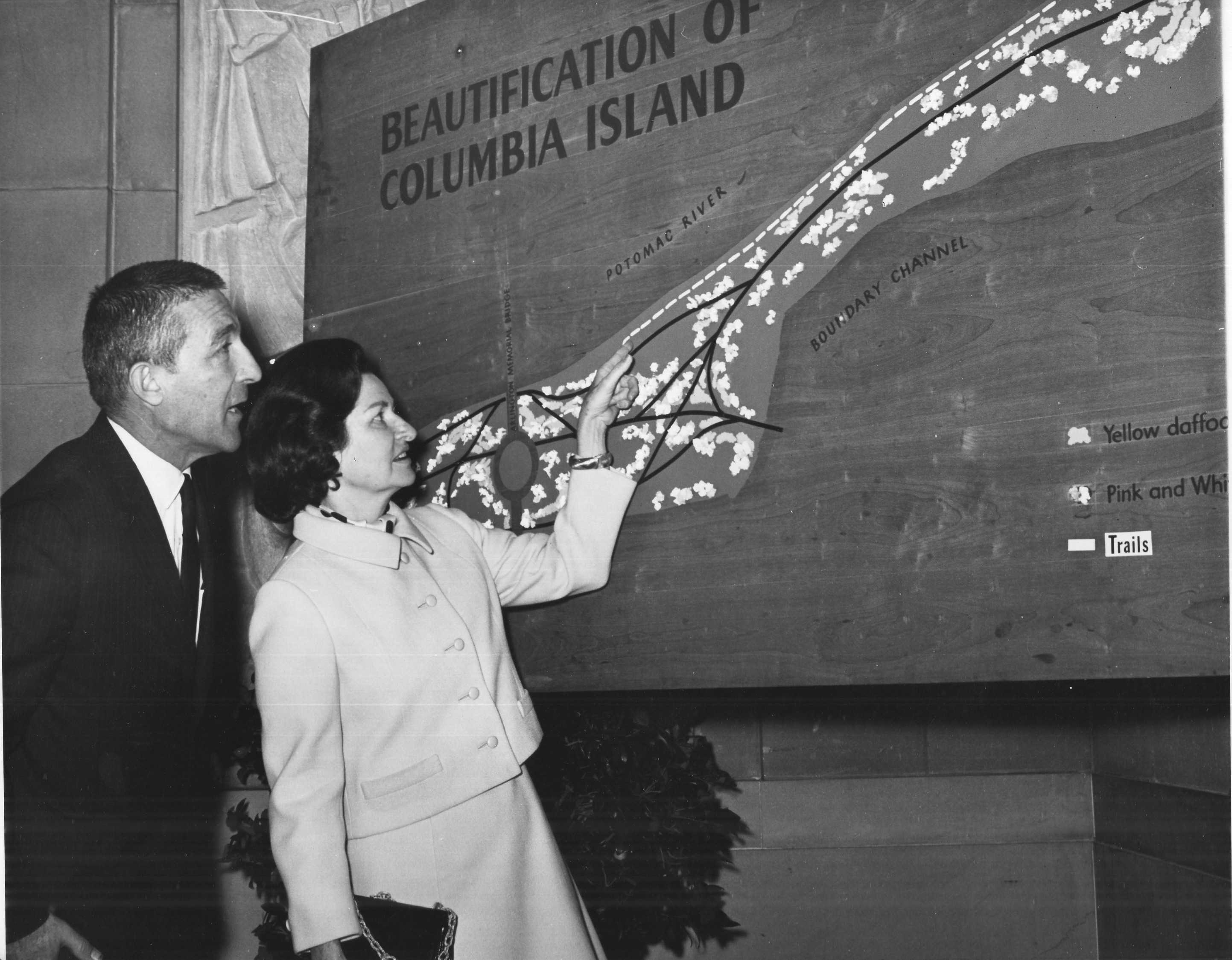 Black and white photo of a well-dressed man and woman pointing to a detail in a large wooden park plan sign, which shows the trails and plants of Columbia Island. 