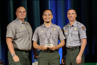 A man holds an award with two men in gray and green uniforms at an award ceremony.
