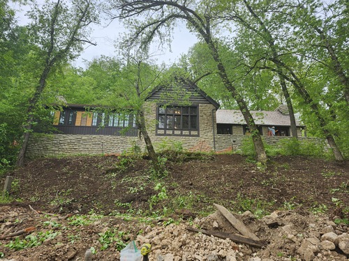 A stone building with a peaked roof, with a center section facing forward and two wings to either side. The left wing has a wooden face and many windows and the right wing is stone with windows, doors, and a porch area. The building is built up on a hillside, seen from below. The hillside has several tall trees with bare earth between them where underbrush has been removed.