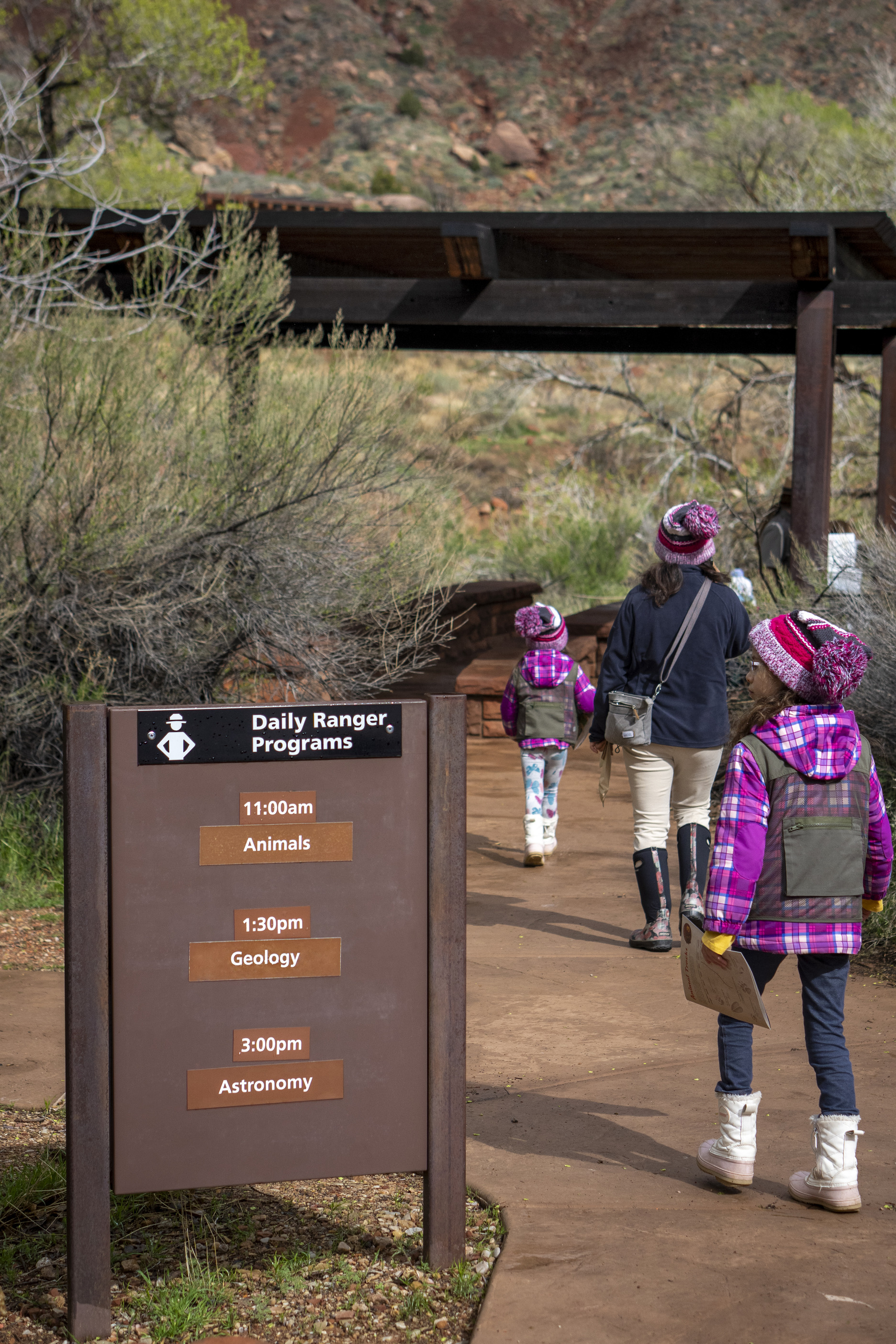 A family of two kids and their mom walk towards a shaded patio for a ranger program. All three are dressed in boots, warm jackets, and beanies. 