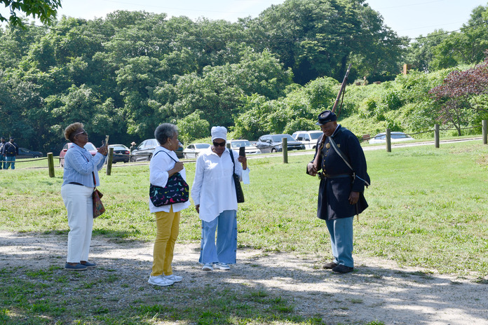 Black man wearing a Union Civil War uniform greets three black women wearing long pant and long sleeve shirts. Two women have their phones our taking his picture.