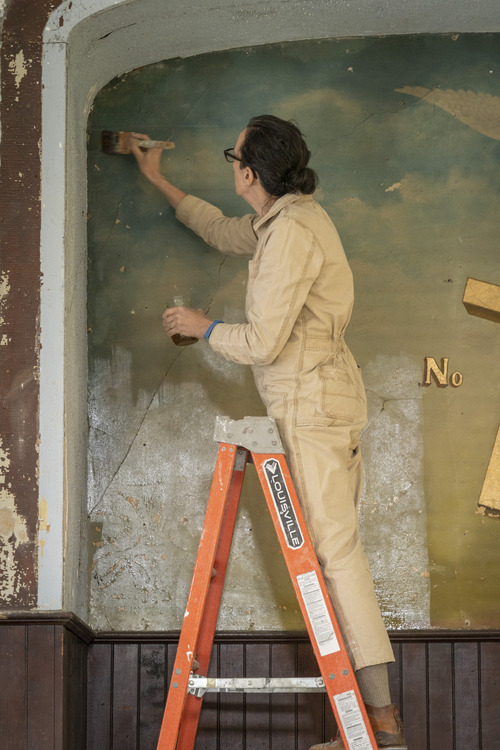 A woman in coveralls stands on a ladder and paints a clear coat on a wall mural.