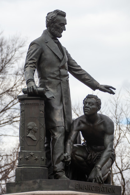 A bronze statue of Abraham Lincoln standing next to a pillar with the Emancipation Proclamation in his hand. His other hand hovers over Archer Alexander, who is kneeling at the feet of Lincoln. Alexander has broken chains around his wrists. 