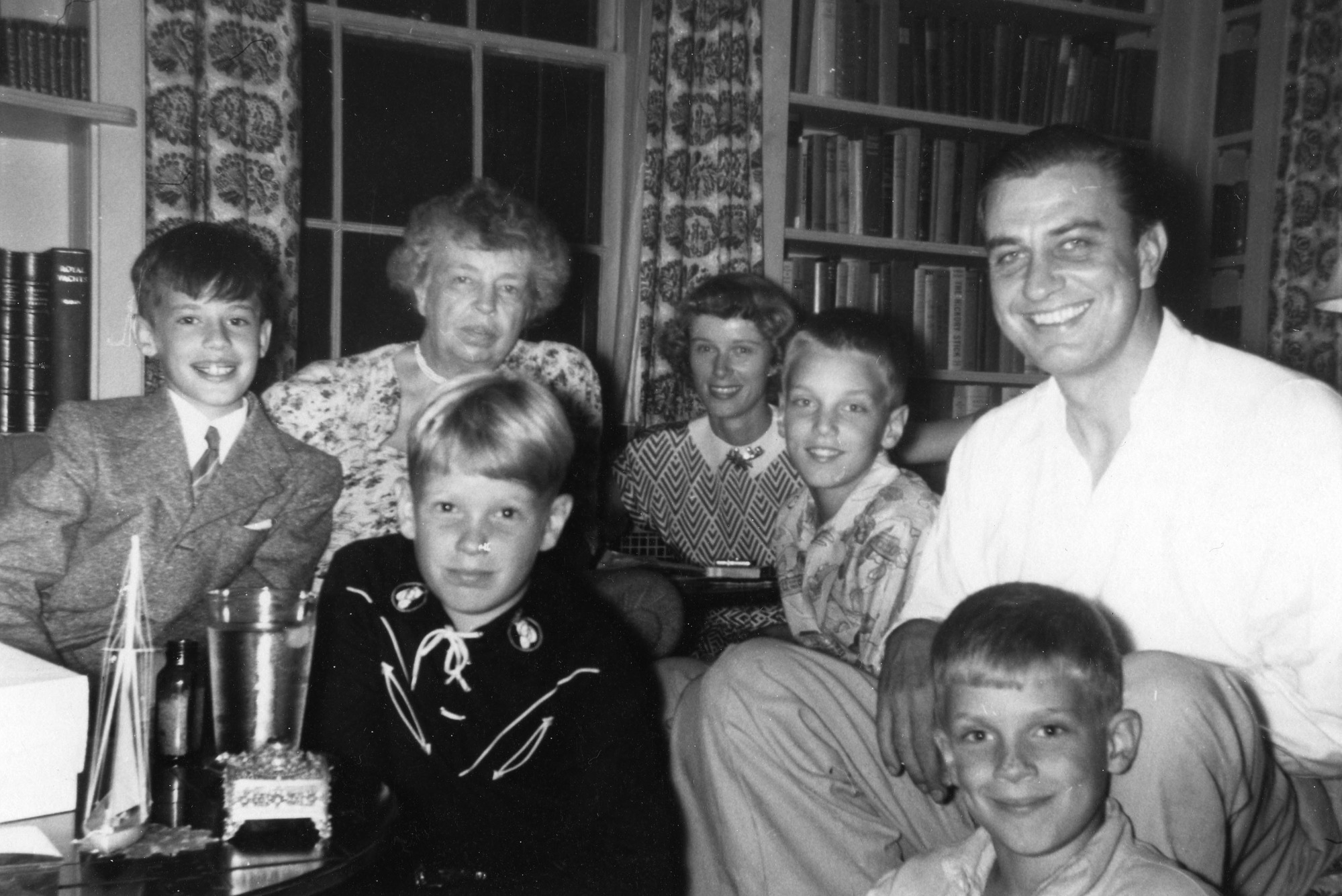 A group photo of four children and three adults in front of a window and bookcases. 