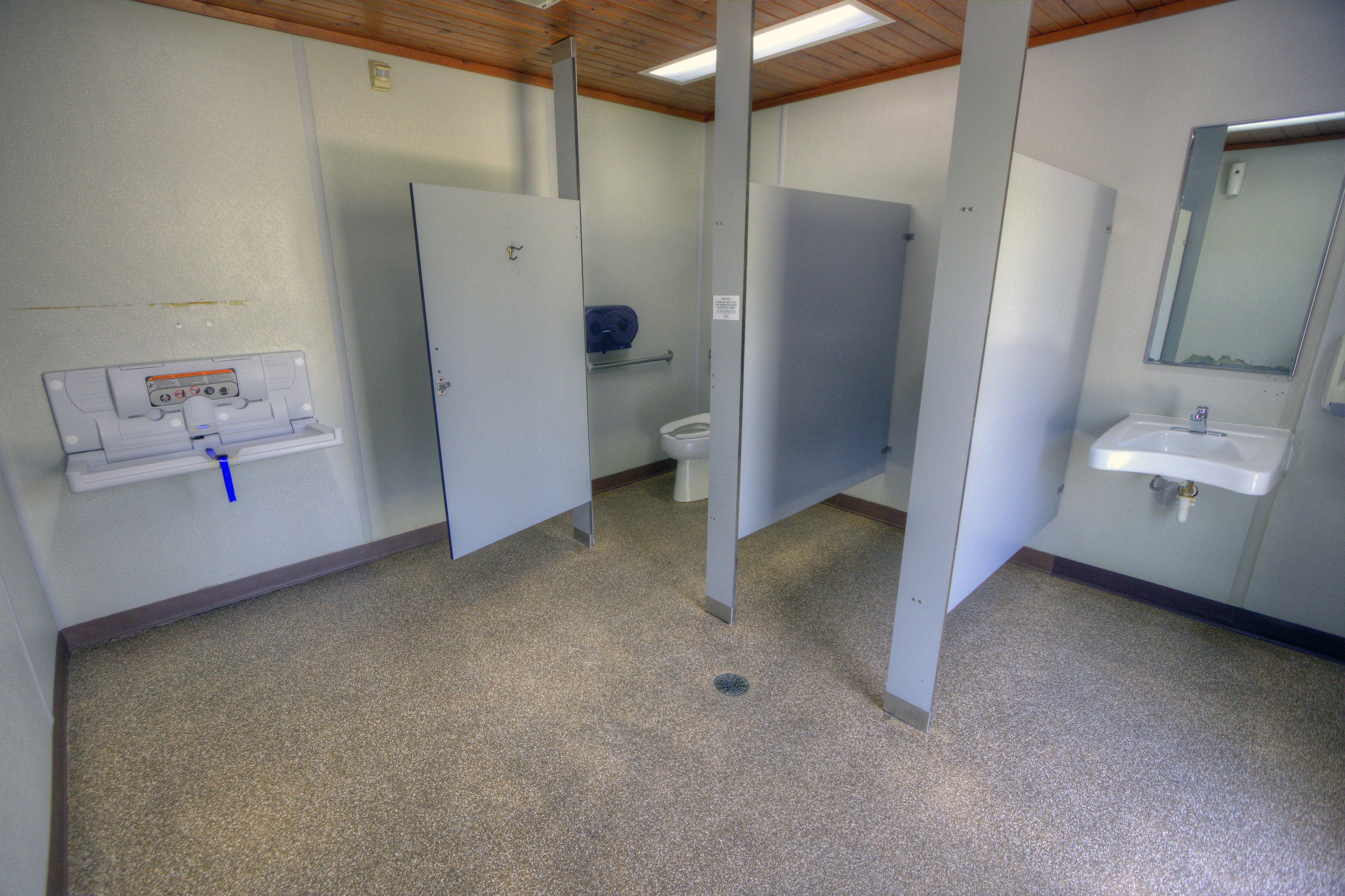 Interior of a restroom. A stall at the far left corner, with a urinal stall and a sink along the same wall. In the far left corner, a changing station lies open.