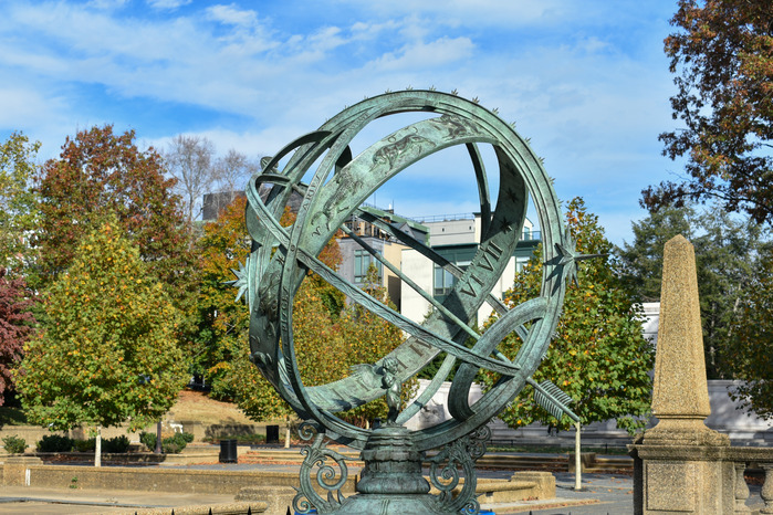 A green-tinted armillary sphere sculpture in front of fall trees and buildings in the distance.
