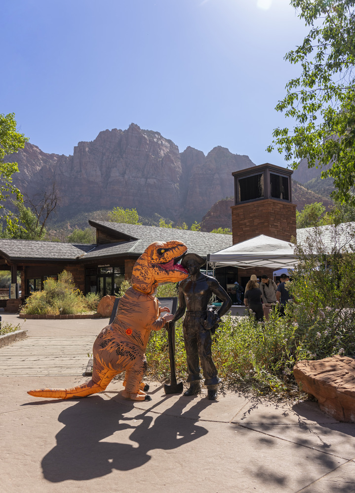 A person in an orange T-Rex costume poses next to a dark statue of a man standing in front of the Zion Canyon Visitor Center. 