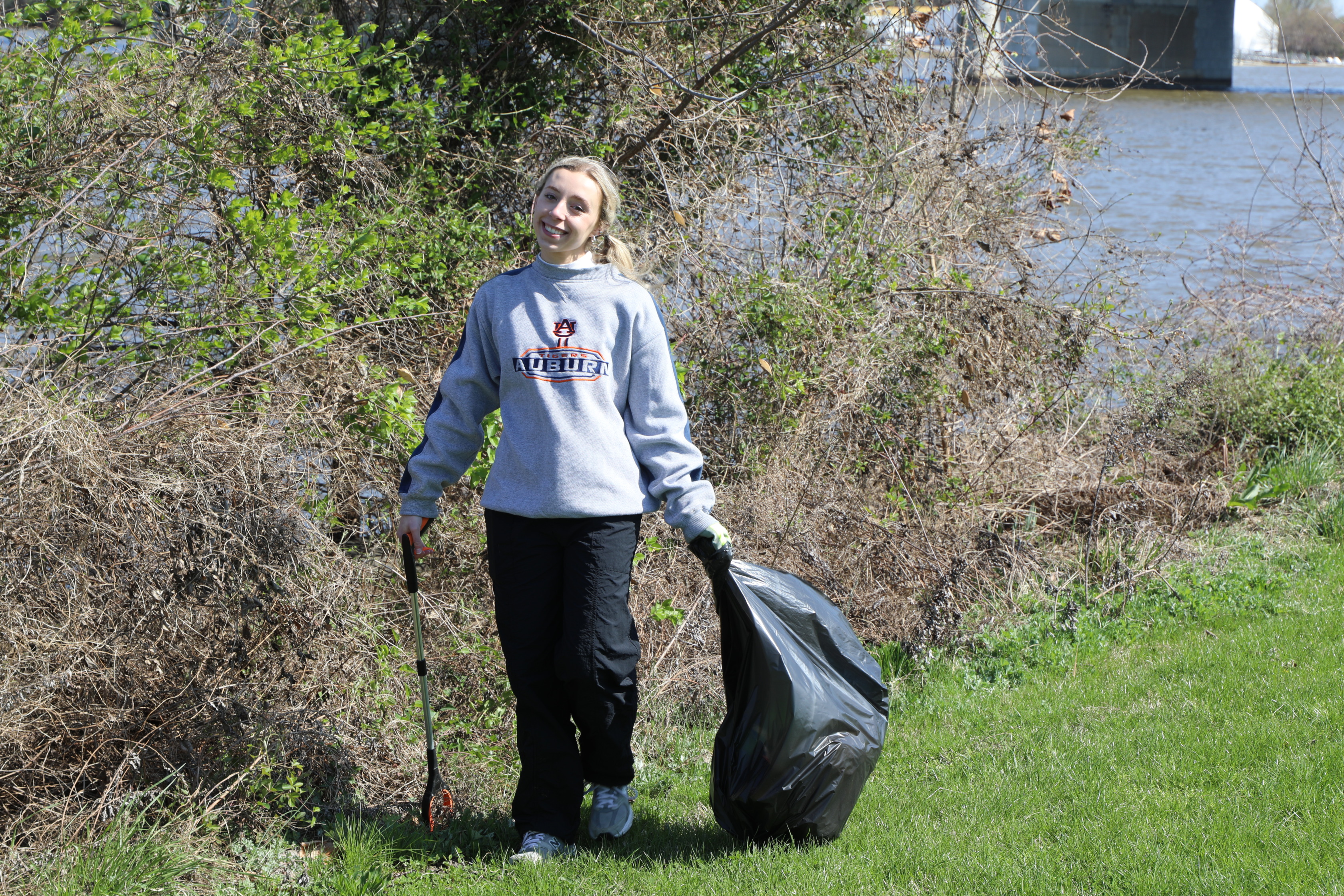 A young woman with blonde hair, wearing a light grey long-sleeved shirt with "AUBURN" on it, black pants, and white sneakers, smiles at the camera. She is standing outdoors in a grassy area, holding a trash grabber tool in her right hand and a black trash bag in her left. Behind her are overgrown bushes, and in the distance, a body of water and a bridge are visible under a bright sky.