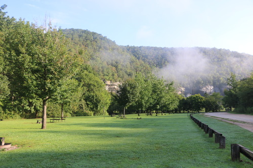 A large green field with fire rings is pictured. In the backdrop is a foggy bluff line.