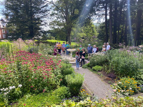 Park employee in red shirt gives tour to large group of visitors standing in the middle of a four square formal garden