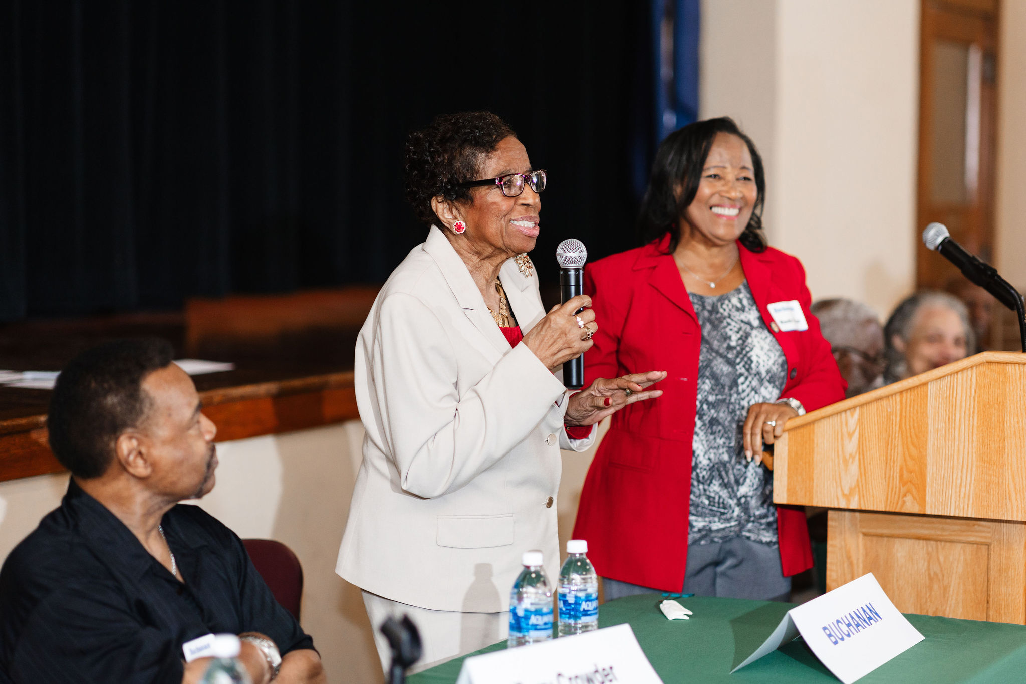 An older african american woman in a white suit speaks into a microphone while standing behind a green table. An african american woman wearing a bright red blazer smiles brightly as she leans against a wooden lectern listening to the first woman
