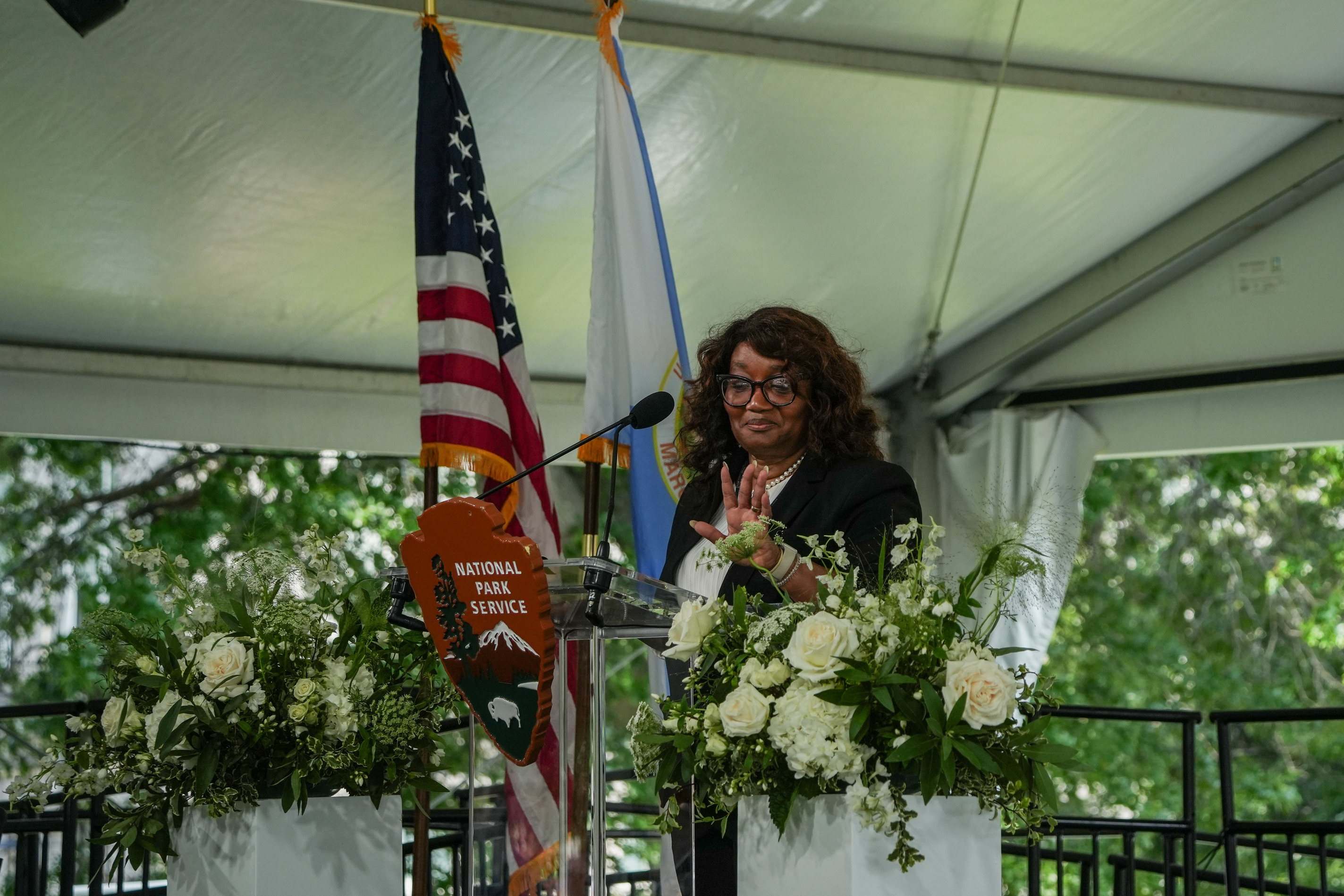 A woman speaks into a microphone at a clear podium. A National Park Service arrowhead hangs from the front of the podium and an US flag and Department of Interior Flag. Flowers are on either side of the podium. 