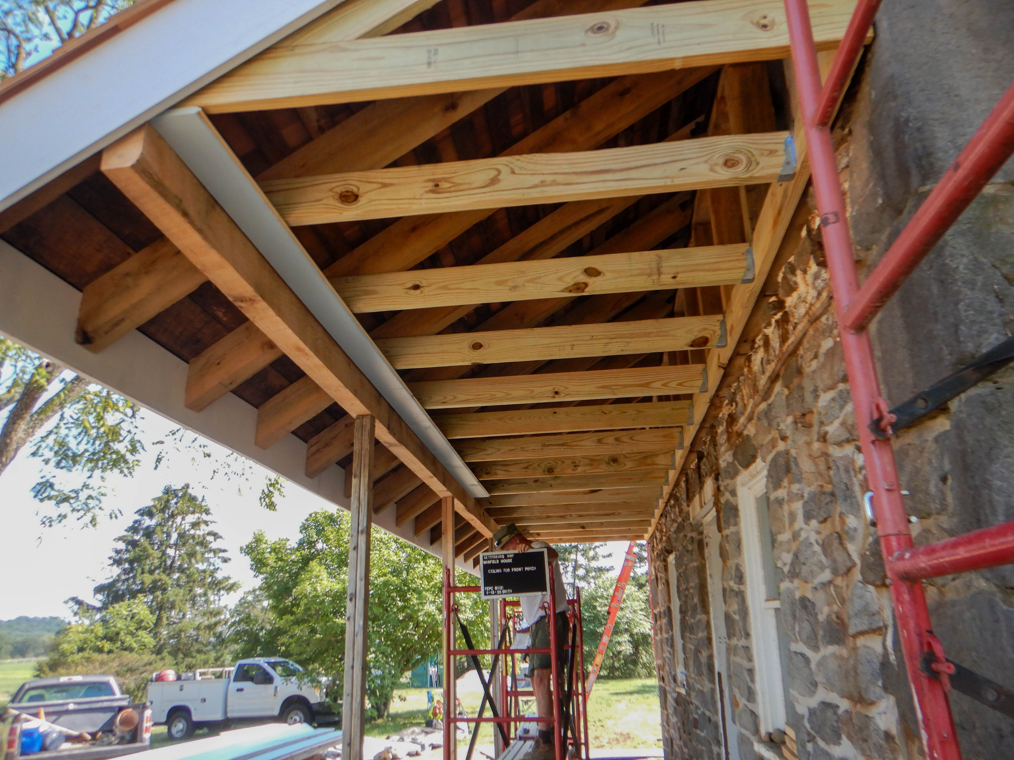 The porch roof ceiling is being constructed. There are ceiling joists in place, which looks like wood framing before the ceiling boards are put into place. A member of the preservation crew stands on a red scaffold and holds and black and white sign that reads "Gettysburg NMP Warfield House Ceiling for front porch PEPC 85531 8-18-20 Smith." There are two trucks full of equipment and trees in the background. 