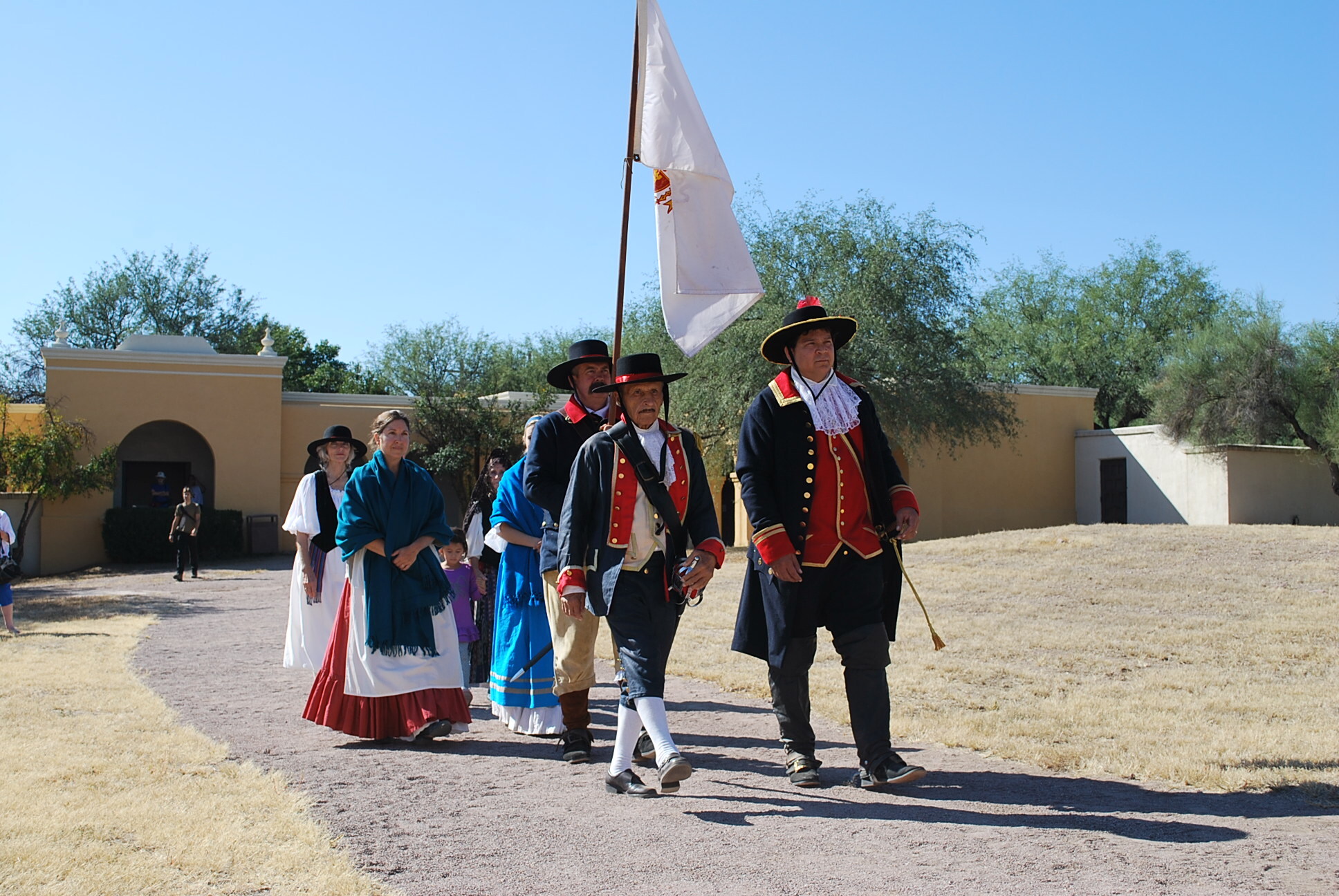 A group of men and women wearing black hats and Spanish colonial dress carry a flag 