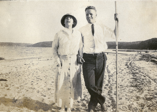 Black and white photograph of man and woman standing next to each other, arms linked on a beach. The woman is wearing a long sleeved blouse, a long skirt, and a brimmed hat. The man is wearing a button down shirt, tie, and trousers. He holds a long pole. They are smiling at the camera. 