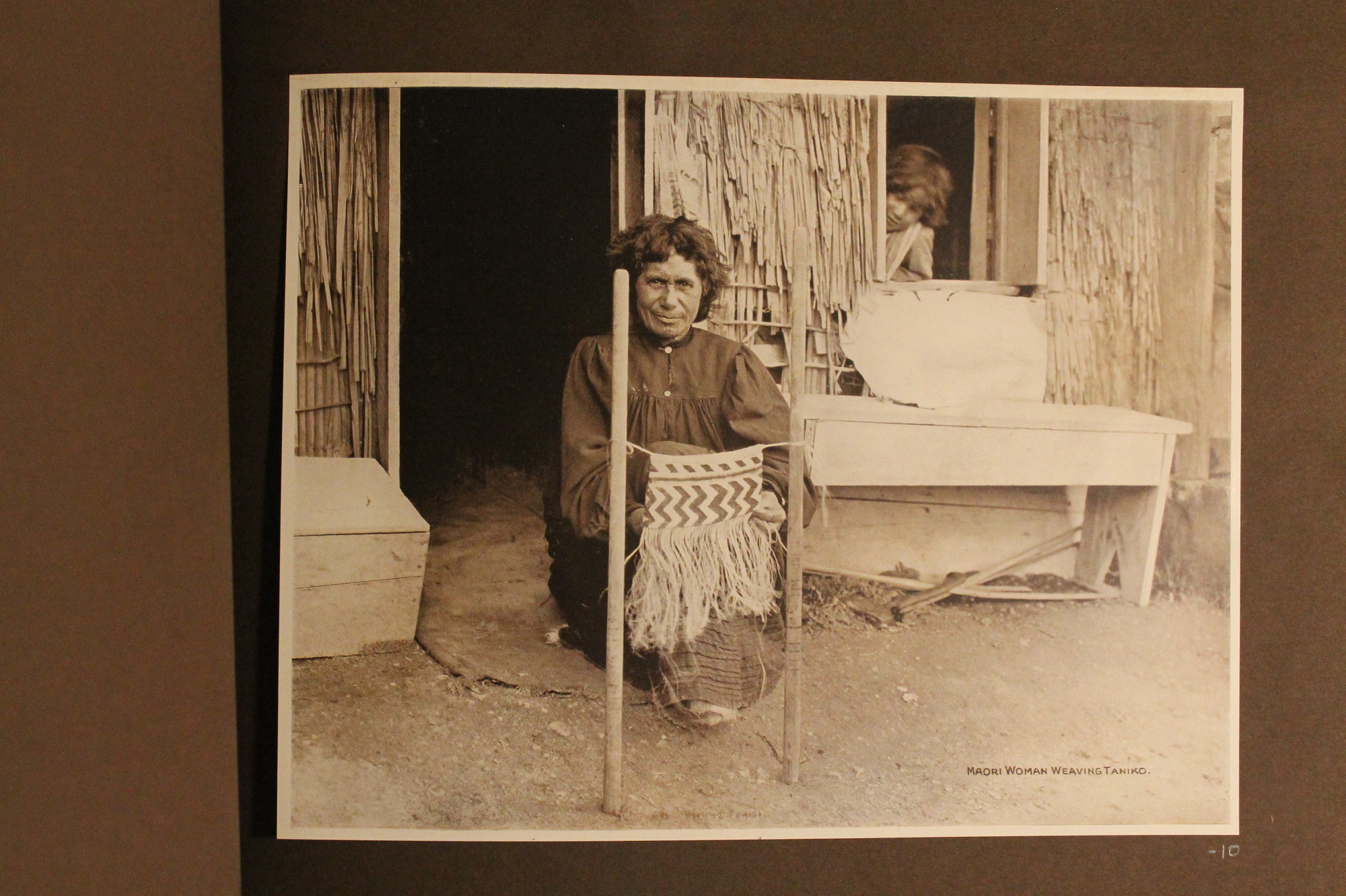 Maori woman weaving in front of straw home.