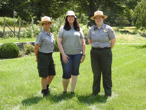 Two Park Rangers standing on either side of an educator wearing Teacher-Ranger-Teacher uniform in on the lawn of a garden at Weir Farm NHP. 