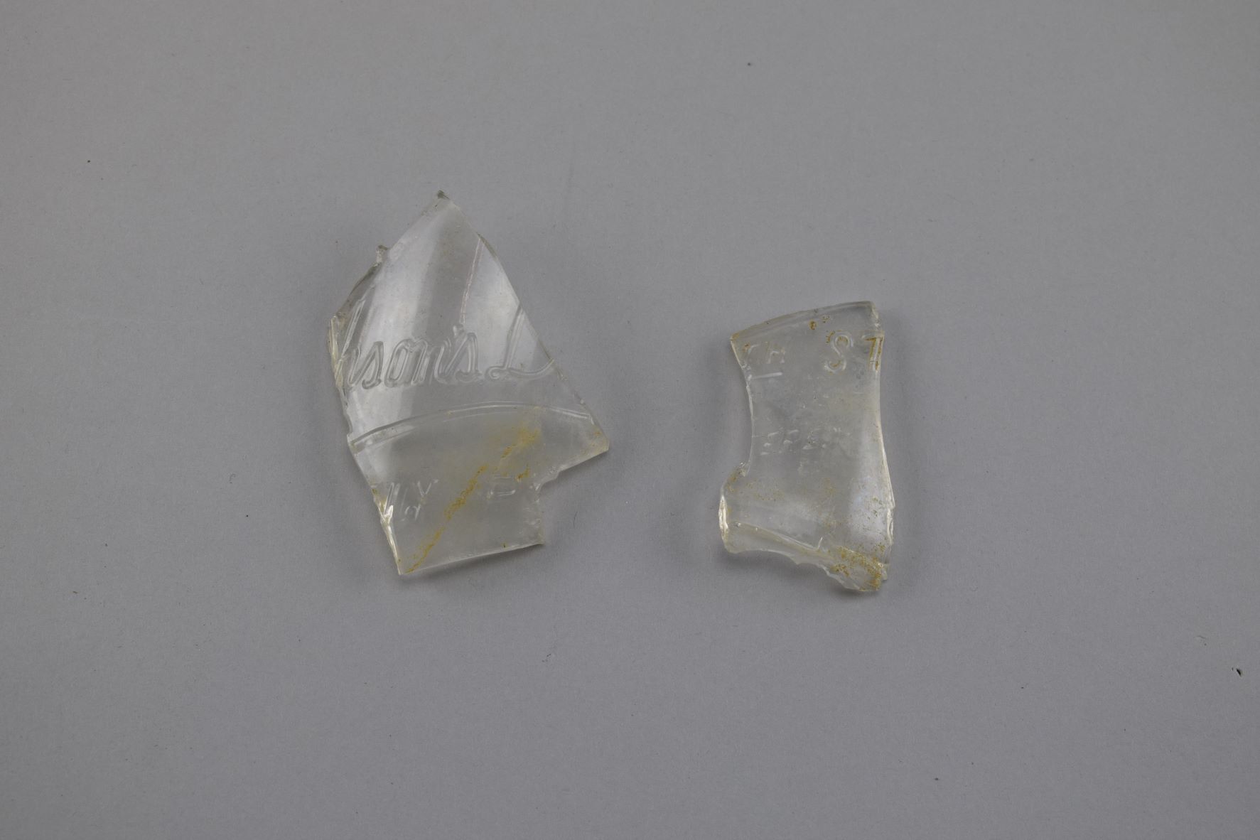 Two small fragments of a clear glass milk bottle. One fragment is embossed with “pson’s D” in cursive and underlined, followed beneath by “LY P”, which were once parts of complete words. The other fragment contains the letters “th St” and “ered”, which were once parts of complete words. 