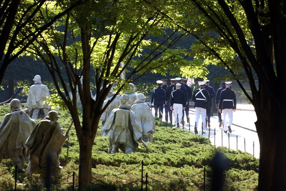 Service members are seen through the trees and shot with backs facing the camera, at left are the statues of soldiers at the Korean War Memorial.