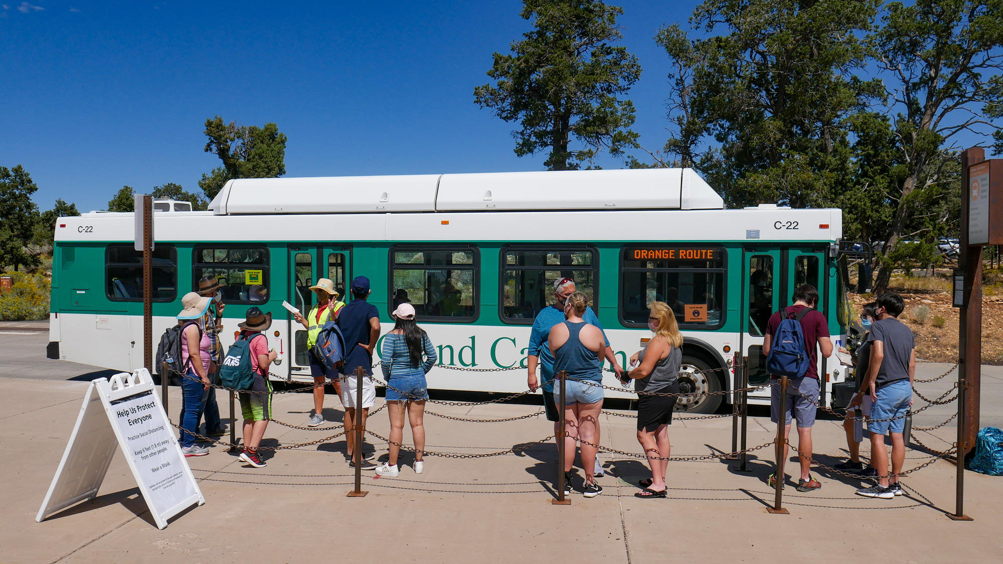 Side view of a large bus with about a dozen people waiting in a line for boarding. 